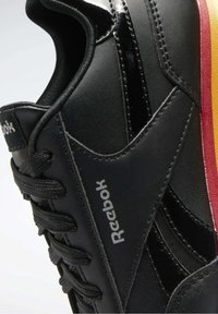 Basket noire Reebok avec lacets noirs, logo blanc sur le côté, et une bande rouge et jaune sur la semelle près du talon.