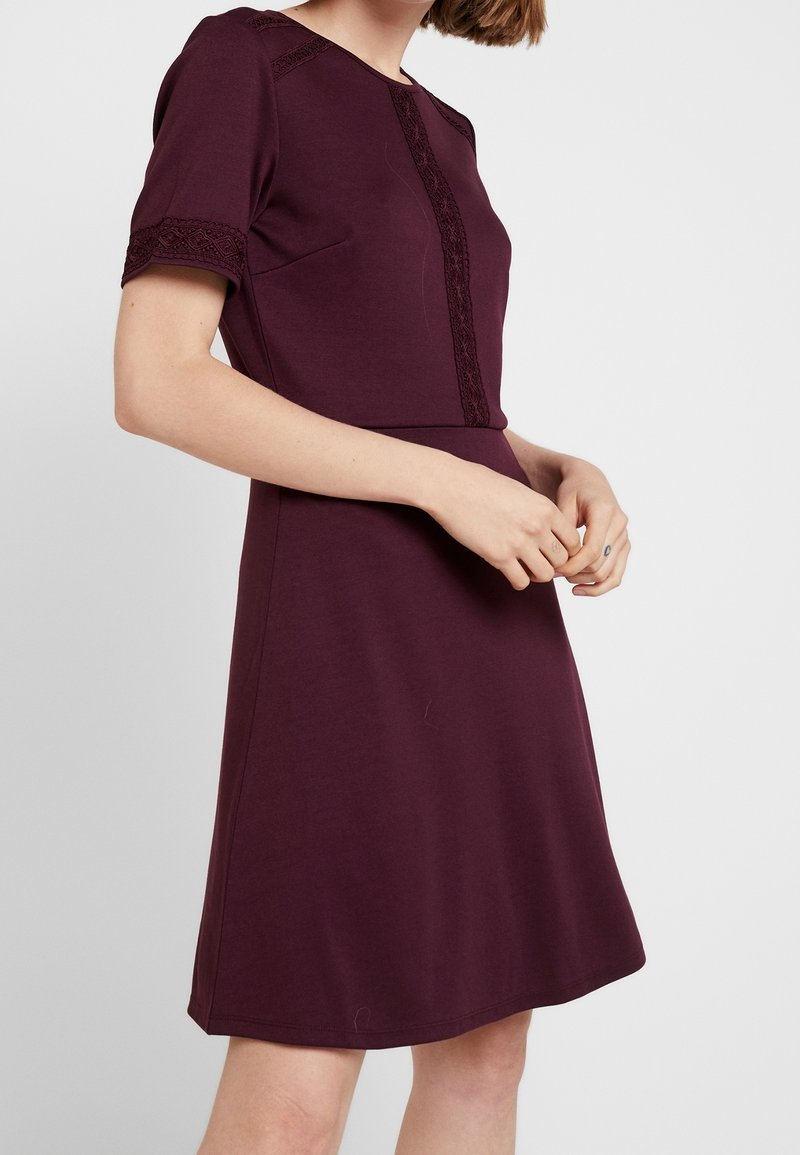 Femme portant une robe prune à manches courtes, longueur genoux, avec une bordure en dentelle aux manches, aux épaules et sur le panneau frontal vertical, debout devant un fond uni.