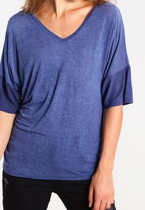 Femme portant une chemise bleue à manches courtes et col en V, ample, avec un pantalon noir, posant avec une main détendue le long du corps.