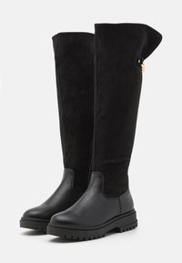 Botas negras hasta la rodilla, combinando materiales de ante y cuero, con un upper texturizado, suela de goma plana y detalles de hardware en dorado.