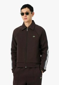 Chaqueta marrón con cremallera, cuello puntiagudo, rayas laterales blancas y logo bordado en el pecho. Tela suave con un diseño ajustado.