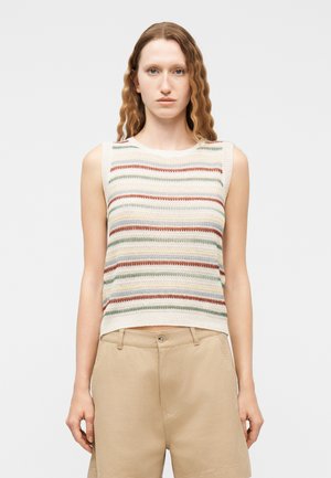 Jeune femme aux cheveux ondulés portant un haut sans manches en tricot à rayures horizontales multicolores et un short beige taille haute.