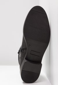 Semelle d'une chaussure en cuir noir avec une prise texturée, taille 37 indiquée, reposant verticalement contre un mur blanc d'angle et le sol.