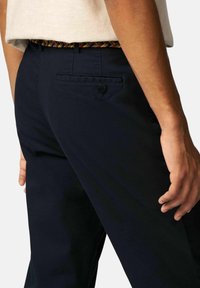 Marineblauwe slim-fit broek met een gladde textuur, voorzien van een achterzak met een knoop, gedragen met een multicolored gevlochten riem.