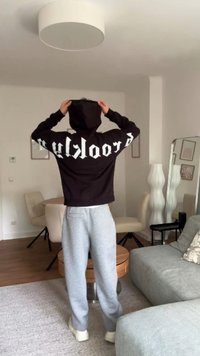 Hanorac negru cu litere albe pe spate, combinat cu pantaloni de trening gri și adidași albi. Setare interioară cu mobilă modernă.