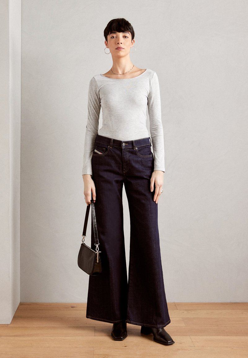 Grauer Langarmoberteil, kombiniert mit dunklen weit geschnittenen Jeans, hält eine kleine schwarze Handtasche. Das Outfit zeichnet sich durch klare Linien und minimales Zubehör aus.