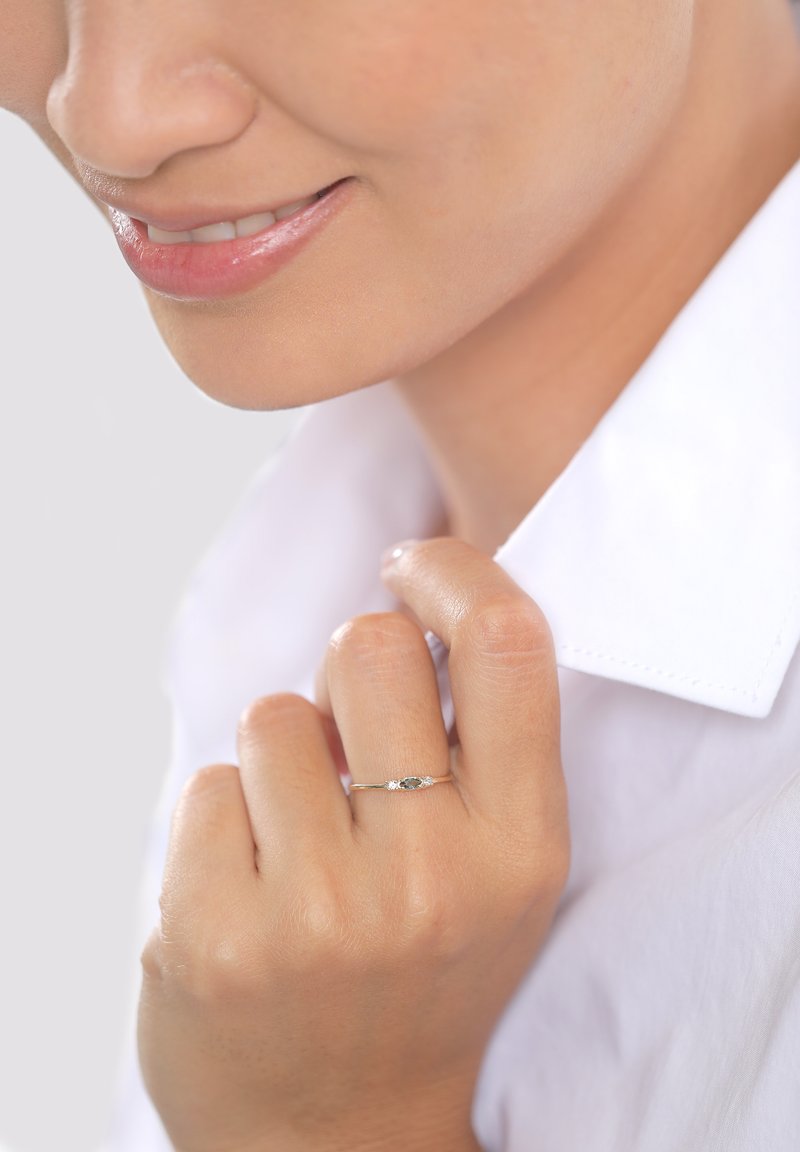 Arany gyűrű ovális kék kővel, két kis áttetsző kővel oldalain; egy fehér galléros ing közelében pihenő kézen viselve.