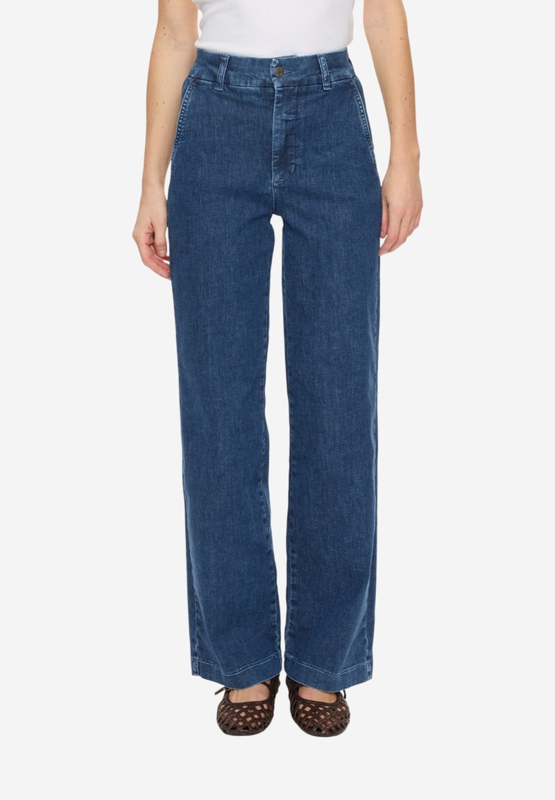 Femme portant un jean bleu taille haute à jambes larges avec poches avant, bouton et fermeture éclair, associé à des chaussures plates tissées marron.