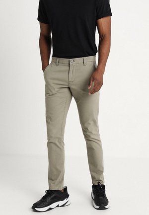 Mężczyzna w beżowych slim fit spodniach, czarnej koszulce i czarnych trampkach, stojący z rękami częściowo w kieszeniach na jednolitym tle.