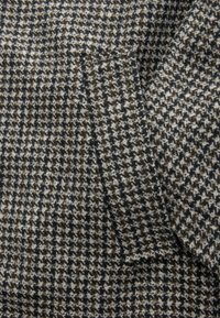 Țesătură din lână cu un model houndstooth în nuanțe de negru, alb și maro, având un mic buzunar inserat. Textură moale, design structurat.