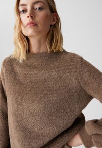 Brauner, flauschiger Pullover mit hohem Kragen und langen Ärmeln; weiche Textur sichtbar; mit entspannter Passform und leichtem Fall der Schultern.