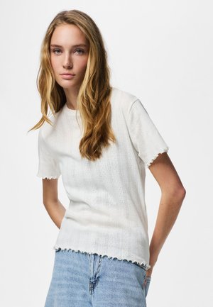 Jauna moteris su ilgais šviesiais plaukais, dėvinti tekstūruotą baltą trumpomis rankovėmis palaidinę ir šviesiai mėlynas džinsines kelnes, stovinti prieš vientisą foną.