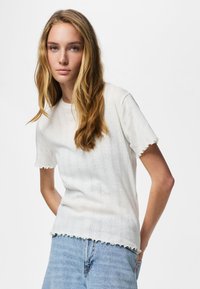 Fiatal nő hosszú szőke hajjal, texturált fehér rövid ujjú felsőt és világoskék farmernadrágot visel, sima háttér előtt áll.