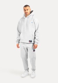 Szara bluza z kapturem z przednią kieszenią i logo na piersi, zestawiona z pasującymi spodniami dresowymi z kieszeniami bocznymi i naszywkami z logo, noszona z białymi sneakersami.
