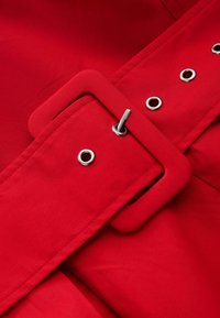 Ceinture en tissu rouge avec boucle rectangulaire recouverte et œillets en métal argenté, passant à travers un œillet, montrant des coutures détaillées.