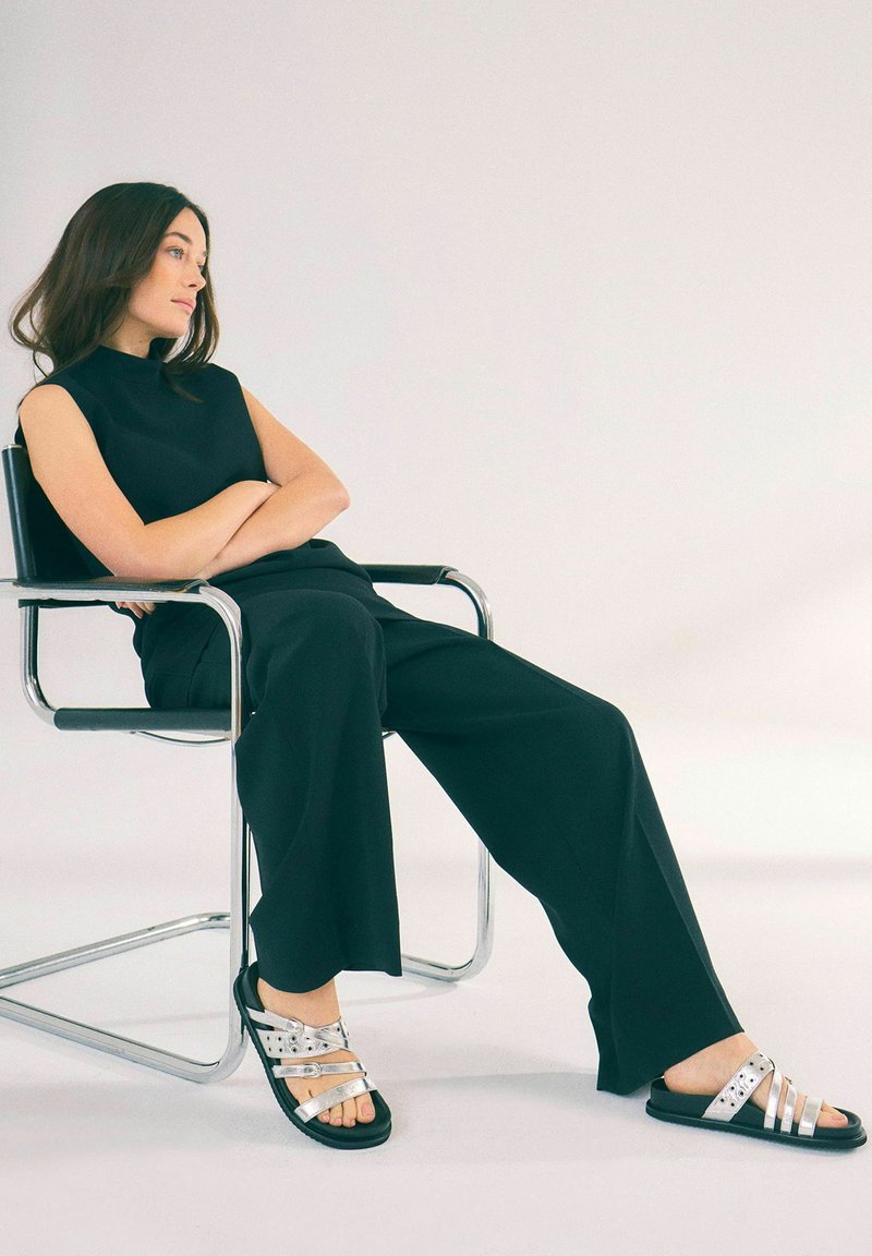 Svart ärmlös topp i kombination med lösa svarta byxor. Modellen bär svarta sandaler med silverremmar och nitar, sittande på en metallstol.