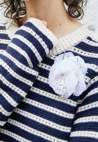 Pull en tricot rayé bleu marine et crème avec un col côtelé. Présente un bijou de fleur en tulle blanc, avec des accents en perles et des épingles en argent.