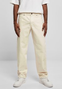Pantalons en denim beige avec une coupe décontractée, dotés d'un design à cinq poches, d'une fermeture à bouton sur le devant et d'une texture subtile. Portés avec des baskets blanches.