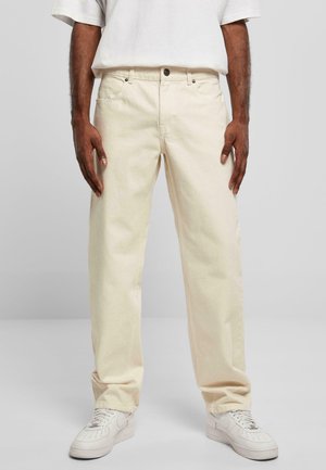 Beige denim pants with a relaxed fit, featuring five-pocket design, front button closure, and subtle texture. Worn with white sneakers.