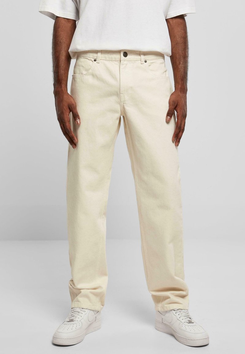 Pantalons en denim beige avec une coupe décontractée, dotés d'un design à cinq poches, d'une fermeture à bouton sur le devant et d'une texture subtile. Portés avec des baskets blanches.