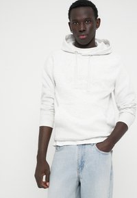 Sweatshirt gris clair avec poche kangourou, capuche à cordon et poignets côtelés. Assorti à un jean bleu clair. Détail de logo subtilement embossé.