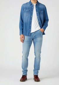 Granatowa koszula dżinsowa z dwiema kieszeniami na piersi, połączona z jasnoniebieskimi dżinsami i brązowymi sznurowanymi butami. Prosty, casualowy design.