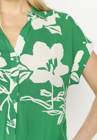Camicia verde con bottoni, realizzata in tessuto morbido, con stampa floreale bianca, maniche corte e colletto. Presenta bottoni verdi coordinati.
