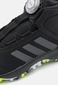 Czarny but sportowy z fakturowaną cholewką, systemem sznurowania BOA i trzema szarymi paskami. Podeszwa z zewnętrzną częścią posiada czarne, białe i zielone kolce.