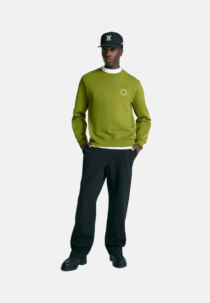 Grüner Pullover mit runder Ausschnitt und kleinem Logo, kombiniert mit schwarzen Hosen und schwarzen Schuhen, getragen von einer Person, die vor einem weißen Hintergrund steht.
