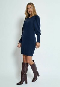 Robe pull en tricot bleu marine avec des poignets et un ourlet côtelés, dotée de manches bouffantes. Associée à des bottes jusqu'aux genoux en vernis bordeaux.