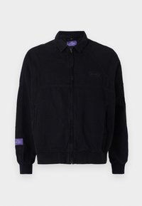 Svart bomberjakke med glidelås, ribbestrikkede mansjetter og krage. Har et brodert logo på brystet og en lilla etikett på ermet.