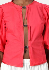 Blusa rossa, corta, con maniche a palloncino, caratterizzata da una chiusura a strappo sul davanti e una texture di tessuto liscia e leggera.