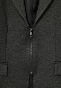 Blazer gris foncé avec une texture lisse, doté d'une fermeture éclair, de deux poches avant et d'une doublure intérieure noire contrastée.
