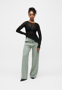 Frau trägt ein langärmliges schwarzes, durchsichtiges Spitzenoberteil, schwarzen BH, salbeigrüne weite Hosen und schwarze spitze Schuhe, steht mit einer Hand in der Hüfte.