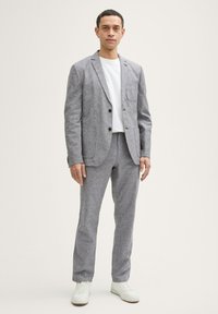 Grau Stoffanzug mit einem einreihigen Blazer und schmal geschnittenen Hosen, kombiniert mit einem weißen T-Shirt und hellen Sneakers. Subtile Textur sichtbar.