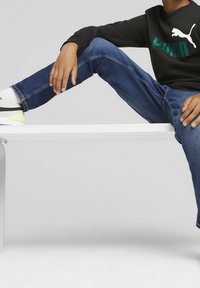 Maglietta nera a maniche lunghe con logo "PUMA" verde, jeans blu a gamba dritta e sneakers bianche con accenti neri e gialli.