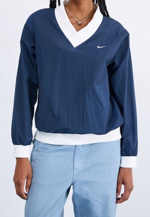 Personne portant un haut bleu marine à manches longues avec un col et des poignets blancs, un jean bleu clair et un petit collier avec pendentif.