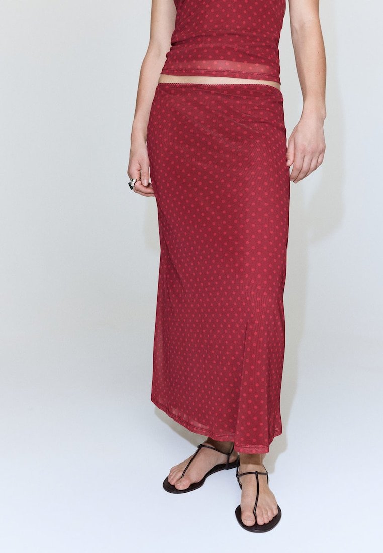 Femme portant une jupe longue à pois rouge et un top sans manches assorti avec des sandales noires à brides, debout devant un fond uni.