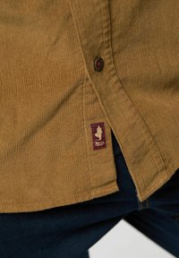 Brun kordfløjlsskjorte med lodrette ribber, udstyret med et maroon label med et logo, sorte knapdetaljer og en lige kant.