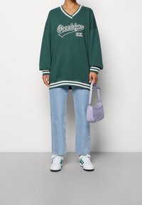 Sweat-shirt vert oversize avec impression Brooklyn blanche, associé à un jean droit bleu clair et des baskets blanches avec des accents verts. Sac violet.