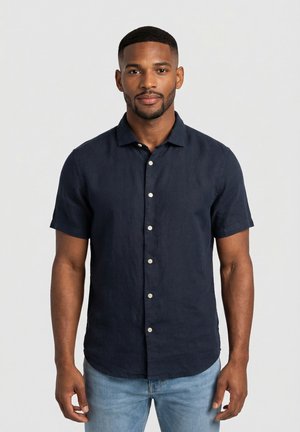 Mężczyzna w granatowej, krótkorękawowej koszuli na guziki i jasnoniebieskich dżinsach, stojący na tle jednolitego białego tła.