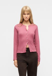 Jeune femme aux longs cheveux blonds portant un cardigan côtelé rose à boutons et un pantalon marron foncé, debout devant un fond uni.