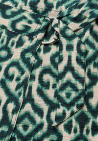 Grönt och krämfärgat mönstrat tyg med abstrakta former. Materialet faller mjukt, med en knuten midjdetalj och metallaccessoarer.