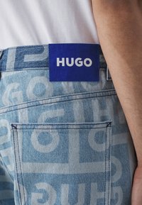 Unos vaqueros de denim azul claro con un patrón de logotipos repetido, y una etiqueta azul en la parte trasera de la cintura que muestra "HUGO" en letras blancas en negrita.