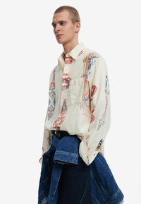 Camicia bianca a maniche lunghe con stampe colorate, taschino frontale e vestibilità comoda. Indossata con una giacca di jeans annodata in vita.