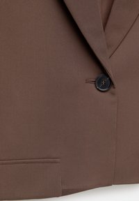 Gros plan d'une veste de costume marron avec un seul bouton noir fermé et des détails visibles sur le revers et la poche.