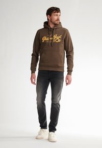 Bruine hoodie met een gele gedrukt logo, kangoeroezak, trekkoorden, gecombineerd met vervaagde zwarte jeans en witte sneakers.