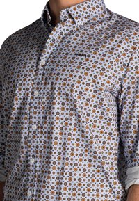 Overhemd met knopen in een lichtblauwe achtergrond en een herhalend bloemenpatroon in oranje, geel en groen. Korte mouwen en kraag.