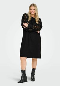 Sort kjole med teksturerte svarte mesh-ermer, løs passform, knelengde, brukt med svarte ankelstøvler. Enkel design med detaljert lagdelt erme.