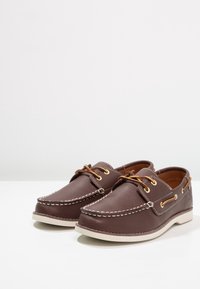 Ein Paar braune Lederboots mit weißer Ziernaht, hellbraunen Schnürsenkeln und weißen Gummisohlen auf einer weißen Fläche.